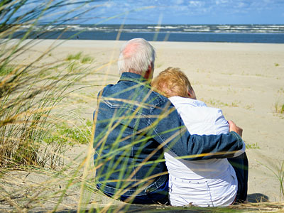 Ein älteres Paar sitzt am Strand und umarmt sich, mit Blick auf das Meer.